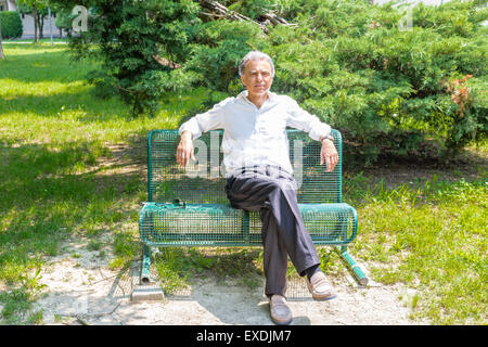 Handsome middle-aged man with salt pepper hair dressed with white shirt, blue slacks and beige moccasins is resting on a bench in city park keeping his arms opened: he shows a reassuring look Stock Photo
