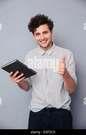 Happy young man holding book and showing thumb up over gray background Stock Photo