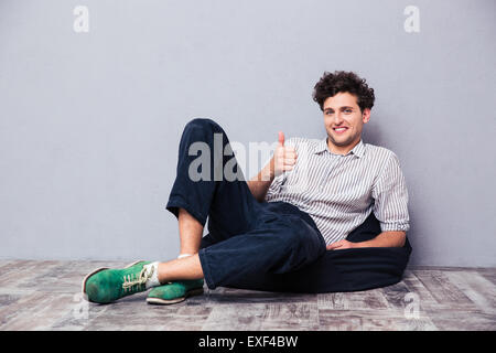 Smiling man sitting on chair bag and showing thumb up over gray background Stock Photo