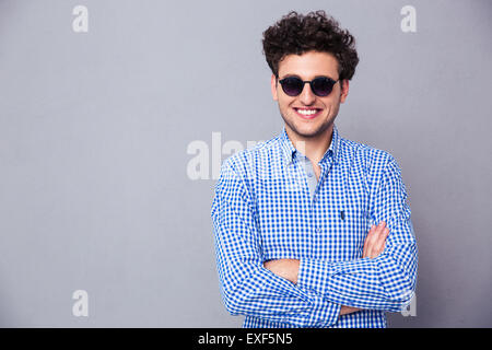 Happy fashion man wearing in sunglasses standing with arms folded over gray background Stock Photo