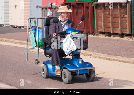 Elderly man on mobility scooter driving past row of beach huts on promenade at Bournemouth in July Stock Photo