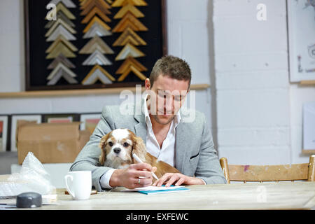 Mid adult man with dog at making notes at desk in picture framers workshop Stock Photo