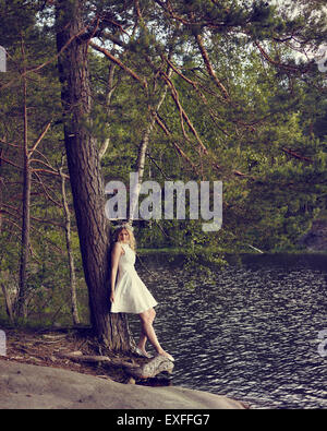 Attractive young woman wearing white summer dress and she leans towards the pine, sunny summer day, lake on background Stock Photo