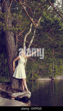 Attractive young woman wearing white summer dress and she leans towards the pine, sunny summer day, lake on background Stock Photo