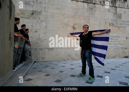 Athens, Greece. 13th July 2015. A protesters poses with a Greek flag next to the Tomb of the Unknown Soldiers in front of riot police officers during the anti-austerity protest outside the Greek Parliament. Greeks assembled outside the Greek Parliament under the banner of 'We leave this Europe'. They called for the government to not give into the demands of the Greek creditors for more austerity measures, but rather to leave the Eurozone. Credit:  Michael Debets/Alamy Live News Stock Photo