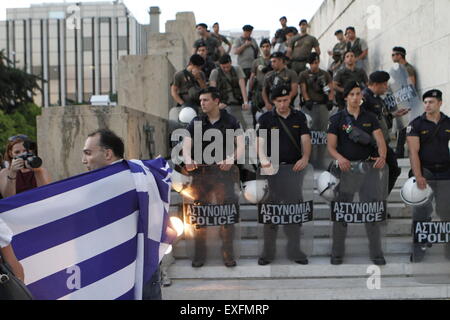 Athens, Greece. 13th July 2015. A protester poses for the press with a Greek flag at the anti-austerity protest on the steps leading up to the Greek Parliament. Riot police officers have secured the steps and watch him. Greeks assembled outside the Greek Parliament under the banner of 'We leave this Europe'. They called for the government to not give into the demands of the Greek creditors for more austerity measures, but rather to leave the Eurozone. Credit:  Michael Debets/Alamy Live News Stock Photo