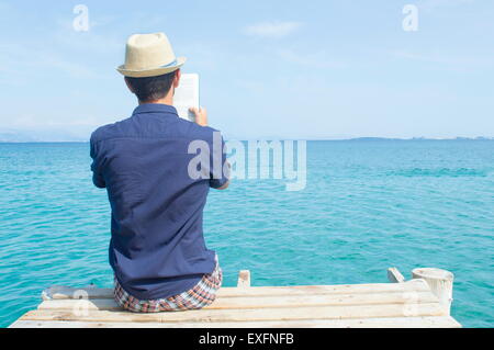 Young man in blue shirt sitting on the dock reading a book Stock Photo