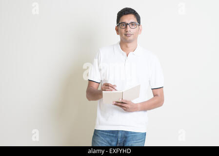 Portrait of Indian guy taking note on booklet. Asian man standing on plain background with shadow and copy space. Stock Photo