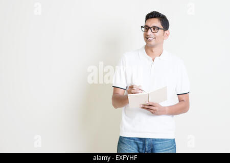 Portrait of Indian guy taking note on book, looking at side and smiling. Asian man standing on plain background with shadow and Stock Photo