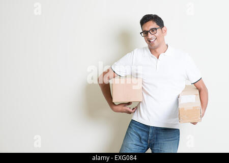 Courier delivery service concept. Happy Indian man holding brown boxes, standing on plain background with shadow. Asian handsome Stock Photo