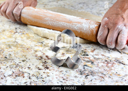 Closeup of a Gingerbread man cookie cutter on a granite counter. In the background are the hands of a baker rolling out dough. Stock Photo