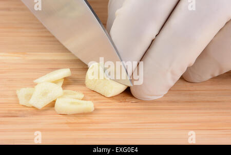 Closeup of a food service worker slicing a garlic clove on a wood cutting board. The chef is wearing a protective glove. Stock Photo