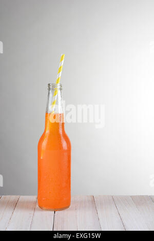 A single bottle of orange soda on a wood table against a light to dark gray background. The bottle has a yellow and white straw Stock Photo