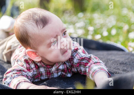 Cute 6 months old baby with Light brown hair in red checkered shirt and beige pants shows a sadly desperate look but fanny face Stock Photo