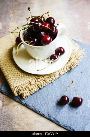 Sweet cherries in a white cup on a wooden table with two cherries in the front Stock Photo