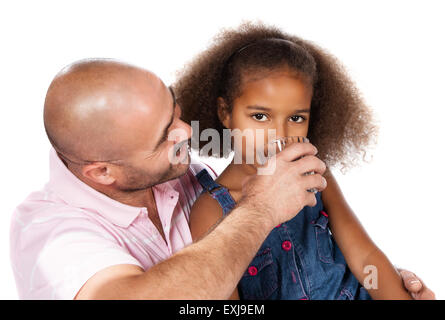 Adorable cute african child with afro hair wearing a denim dress. Her father is helping her to drink water from a clear glass. Stock Photo