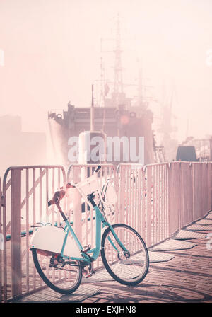 Vintage toned bicycle parked by pier with ship in a distance, sunset lens flare effect. Stock Photo