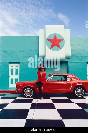 Lady wearing red posing with a red Mustang car, in a very colorful setting. Stock Photo