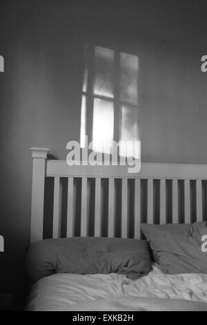 Unmade double bed in bedroom showing headboard and pillows, shadow of window on wall above, in black and white film noir style Stock Photo