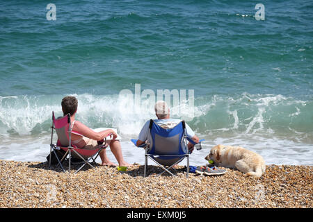 Torcross, Devon, UK. Two people and dog sat in beach chairs at water's edge on shingle beach at Torcross in Devon. Stock Photo
