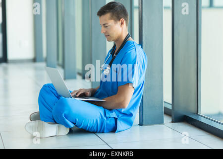handsome medical doctor sitting on hospital floor and using laptop Stock Photo