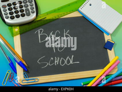 Back to school message written in chalk on blackboard surrounded by school or office supplies on a blue green background Stock Photo