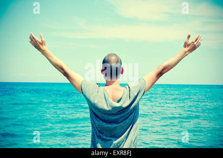 closeup of a young caucasian man seen from behind with his arms in the air in front of the ocean, feeling free, with a slight vi Stock Photo
