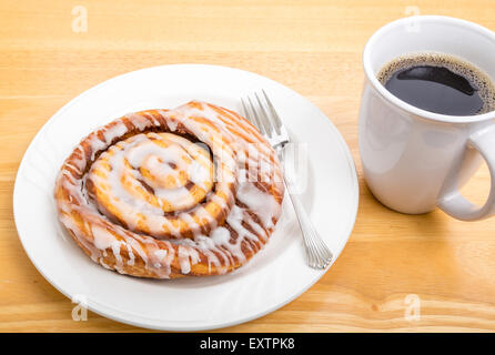 A fresh cinnamon roll on a plate with fork and a cup of coffee Stock Photo