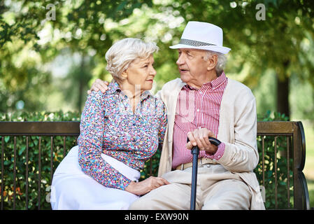 Restful seniors in smart casual sitting on bench in park and talking Stock Photo