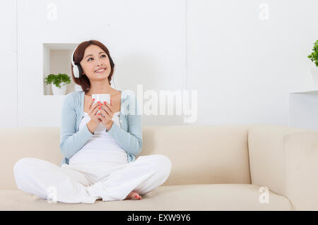 Young woman sitting on sofa, holding a cup and wearing headphone, Stock Photo