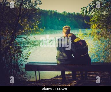 Silhouettes of hugging couple sitting on bench against the lake at sunset. Vintage photo. Stock Photo