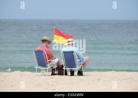 St Ives, Cornwall, UK: Two Middle-aged men wearing jumpers and straw hats sat on the beach on camping chairs. Stock Photo