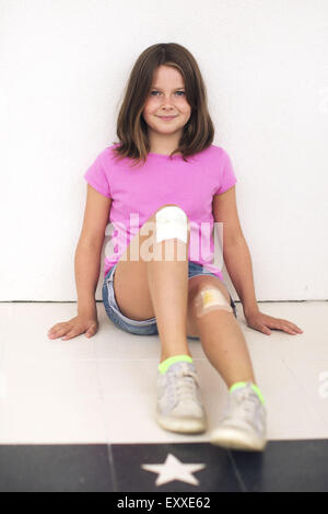 Girl sitting on floor with both knees bandaged, smiling Stock Photo