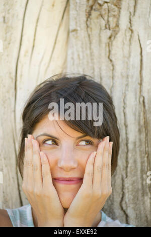 Woman leaning against tree trunk, smiling and holding face in hands, portrait Stock Photo