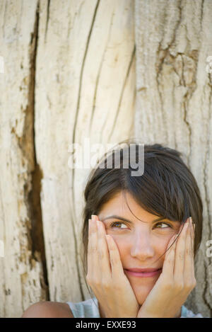 Woman leaning against tree trunk, smiling and holding face in hands, portrait Stock Photo