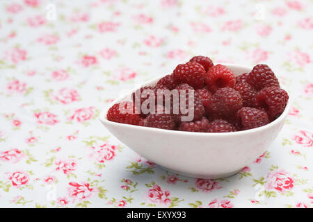 Rubus idaeus 'Autumn Bliss'. Freshly picked red berries in a bowl on a flowery cloth. Stock Photo