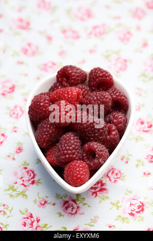 Rubus idaeus 'Autumn Bliss'. Freshly picked red berries in a bowl flowery cloth. Stock Photo