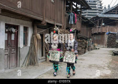 Dong woman in traditional attire walking in Huanggang Dong Village with large drum tower, Guizhou Province, China Stock Photo