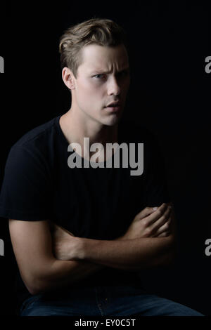 A blond, Caucasian man, sitting with arms crossed. He appears to be frustrated and in disagreement. A dark portrait. Stock Photo
