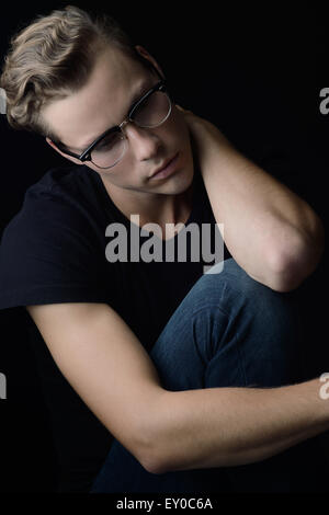 A blond wavy hair man, sitting and looking away with sadness. He appears to be lonely and emotional. A dark portrait. Stock Photo