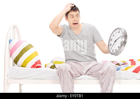 Shocked young man sitting on a bed in his pajamas and looking at the time isolated on white Stock Photo