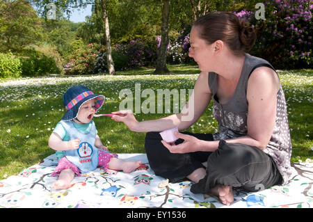 Baby girl wearing a sun hat sitting on a rug with mum being spoon fed yogurt Stock Photo