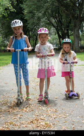 Three children wearing safety helmets and riding scooters in a New York park USA Stock Photo
