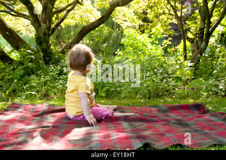 Baby girl sitting unaided on a rug outdoors looking away Stock Photo