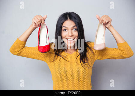 Cheerful young woman holding shoes over gray background and looking at camera Stock Photo