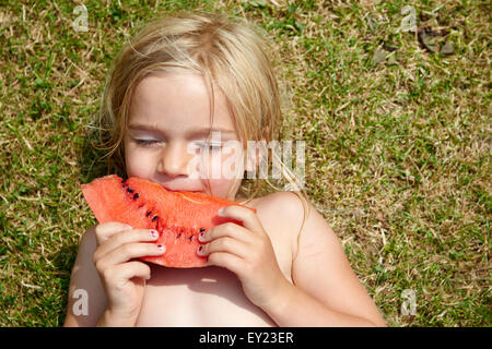 Portrait of little child blond girl with slice of watermelon, lying on green grass lawn, summer, relax Stock Photo