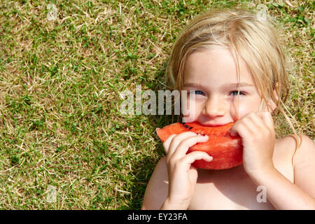 Portrait of little child blond girl eating slice of watermelon, lying on green grass lawn, summer, relax Stock Photo