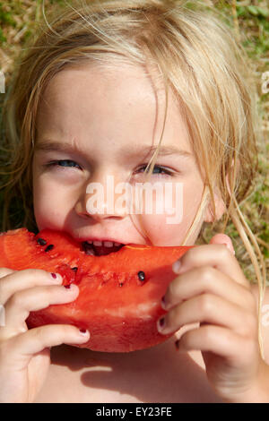 Portrait of little child blond girl eating slice of watermelon, lying on green grass lawn, summer, relax Stock Photo