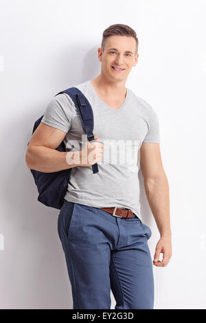 Vertical shot of a casual male student with a blue backpack leaning against a wall and looking at the camera Stock Photo