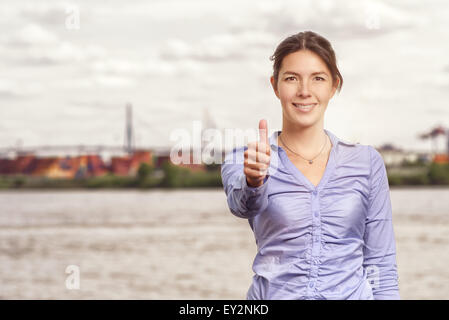 Happy smiling attractive woman giving a thumbs up gesture of success and approval, upper body portrait in front of an urban rive Stock Photo